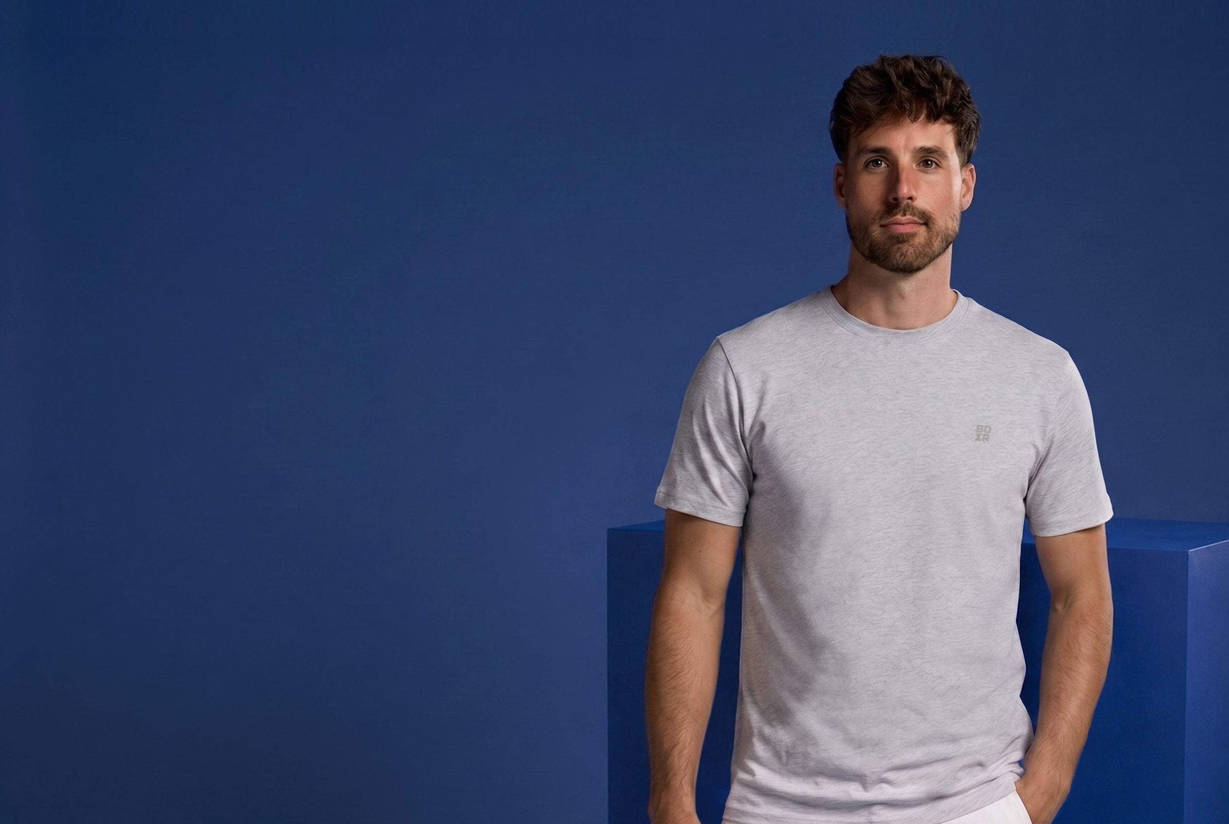A man wearing a light gray crewneck t-shirt stands against a solid blue background.
